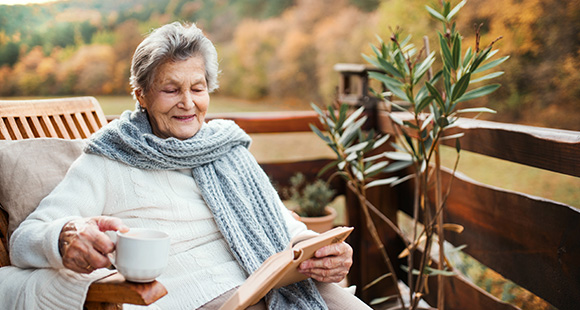 an elderly lady wearing a scarf reading a book outdoors while holding a cup of tea